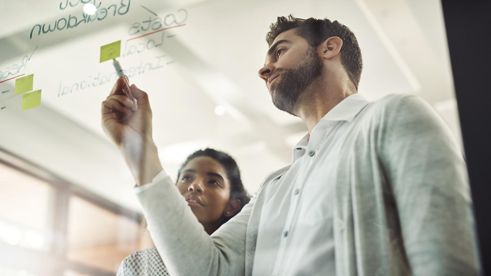 Ein Mann und eine Frau arbeiten mit Post-Its und Filzstift an einer Glasscheibe