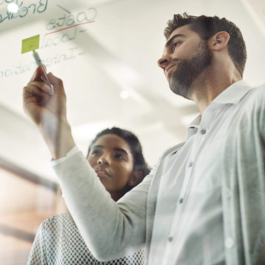 Ein Mann und eine Frau arbeiten mit Post-Its und Filzstift an einer Glasscheibe